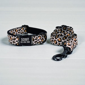 May include: A dog collar and leash set with a leopard print pattern. The collar has a black buckle with the name "SCOUT" and a phone number. The leash is rolled up next to the collar.