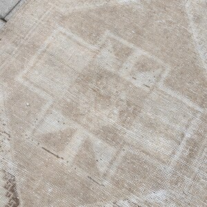 Puede incluir: Una alfombra rectangular de color beige con un aspecto descolorido y desgastado. La alfombra presenta un diseño geométrico central en un tono ligeramente más oscuro, creando un contraste sutil. La alfombra está colocada sobre una superficie gris texturizada.