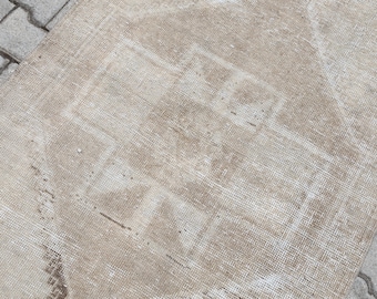 May include: A rectangular, beige-toned rug with a faded, distressed appearance. The rug features a central geometric design in a slightly darker shade, creating a subtle contrast. The rug is placed on a gray, textured surface.