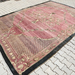 May include: A vintage rug with a pink background and a black and gold border. The rug has a floral pattern and a geometric design in the center.
