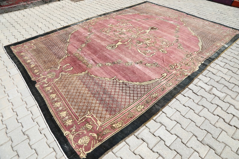 May include: A vintage rug with a pink background and a black and gold border. The rug has a floral pattern and a geometric design in the center.
