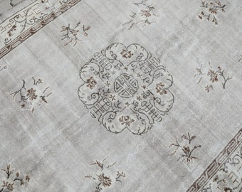 May include: A light gray rug with a brown floral pattern. The rug has a central design with a stylized flower and a square in the center. The rug has a border with a geometric pattern.