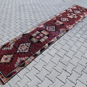 Puede incluir: Una alfombra larga y estrecha de color rojo y crema con un patr&oacute;n geom&eacute;trico. La alfombra tiene un patr&oacute;n de diamantes con acentos negros.