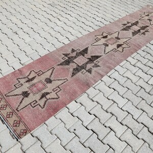 May include: A long, narrow rug with a geometric pattern in shades of pink, brown, and white. The rug is laid out on a gray brick surface.