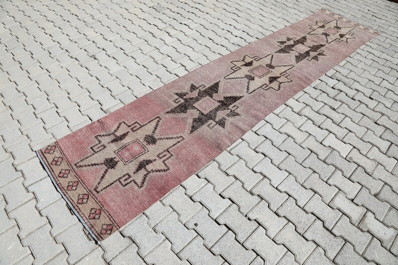 May include: A long, narrow rug with a geometric pattern in shades of pink, brown, and white. The rug is laid out on a gray brick surface.