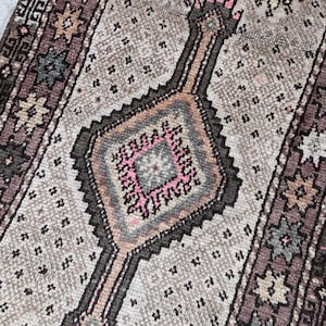 Puede incluir: Una alfombra rectangular de estilo vintage con un diseño central de diamante en tonos rosa, marrón y gris. La alfombra presenta un patrón repetitivo de pequeñas formas negras y motivos florales a lo largo de los bordes. La paleta de colores general es apagada.