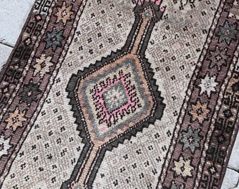 Puede incluir: Una alfombra rectangular de estilo vintage con un diseño central de diamante en tonos rosa, marrón y gris. La alfombra presenta un patrón repetitivo de pequeñas formas negras y motivos florales a lo largo de los bordes. La paleta de colores general es apagada.
