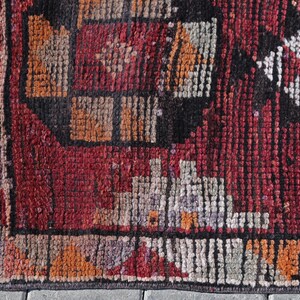 Puede incluir: Primer plano de una alfombra roja, negra y blanca con un borde de flecos. La alfombra tiene un dise&ntilde;o geom&eacute;trico con cuadrados y rect&aacute;ngulos.