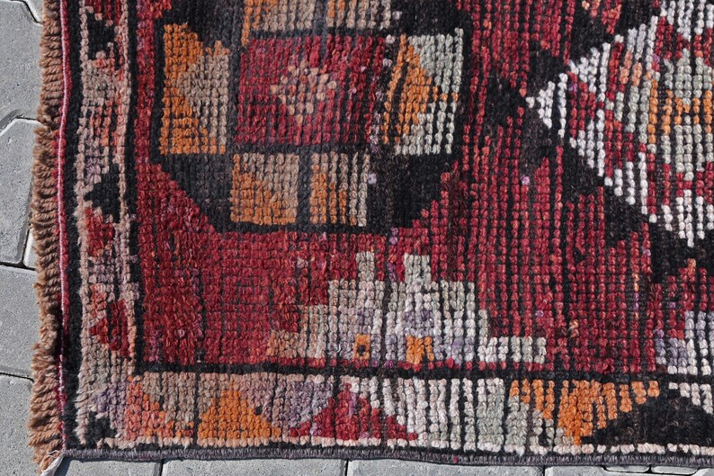 Puede incluir: Primer plano de una alfombra roja, negra y blanca con un borde de flecos. La alfombra tiene un dise&ntilde;o geom&eacute;trico con cuadrados y rect&aacute;ngulos.