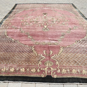 May include: A vintage pink and beige rug with a floral pattern and a black border. The rug has a worn and faded look, giving it a rustic charm.