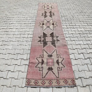 May include: A long, narrow rug with a pink background and a geometric pattern in shades of brown and beige. The rug has a fringe on both ends.