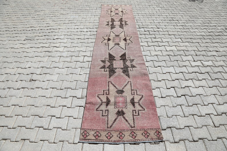 May include: A long, narrow rug with a pink background and a geometric pattern in shades of brown and beige. The rug has a fringe on both ends.