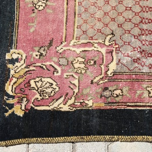 May include: Close-up of a corner of a vintage rug with a black border, a pink and cream floral pattern, and a geometric design in the center.