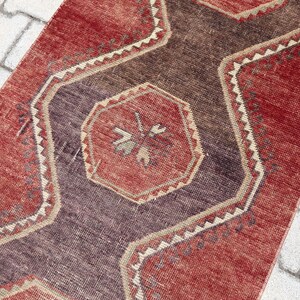 Peut inclure: Un tapis rectangulaire vintage avec un motif géométrique central. Le tapis présente une palette de couleurs rouge, marron et violet, avec une bordure beige. Le motif du tapis comprend des formes octogonales et des motifs floraux.