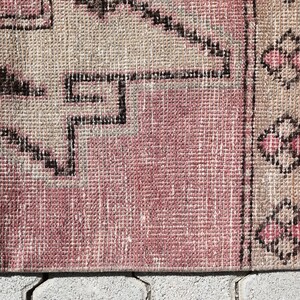 May include: Close-up of a pink and beige woven rug with a geometric pattern. The rug has a fringe border and is made of a soft, textured material.