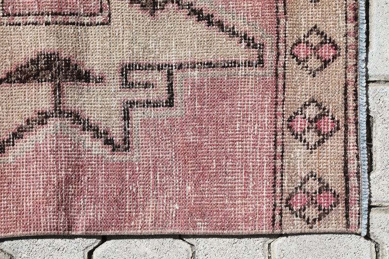 May include: Close-up of a pink and beige woven rug with a geometric pattern. The rug has a fringe border and is made of a soft, textured material.