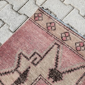 May include: A pink and beige patterned rug with a geometric design. The rug has a frayed edge and is lying on a gray concrete surface.