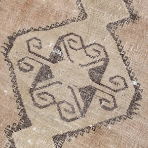 Puede incluir: Una alfombra tejida con un color base bronceado claro y un prominente medallón geométrico marrón oscuro en el centro. El diseño incluye intrincados patrones angulares de color marrón oscuro y pequeños acentos de verde apagado.