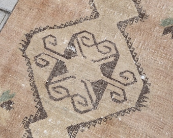 May include: A woven rug featuring a light tan base color with a prominent dark brown geometric medallion in the center. The design includes intricate dark brown angular patterns and small accents of muted green throughout the textile surface.