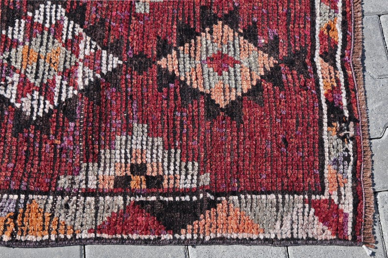 Puede incluir: Primer plano de una alfombra roja, negra y blanca con un borde de flecos. La alfombra tiene un dise&ntilde;o geom&eacute;trico con formas de diamante.