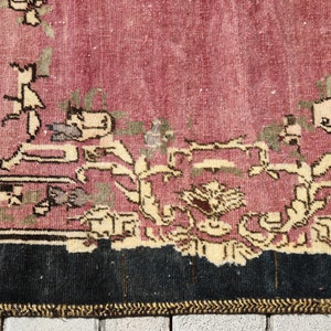 May include: Close-up of the bottom edge of a vintage rug with a pink background and a floral pattern in shades of cream, brown, and green. The rug has a black border and a gold fringe.