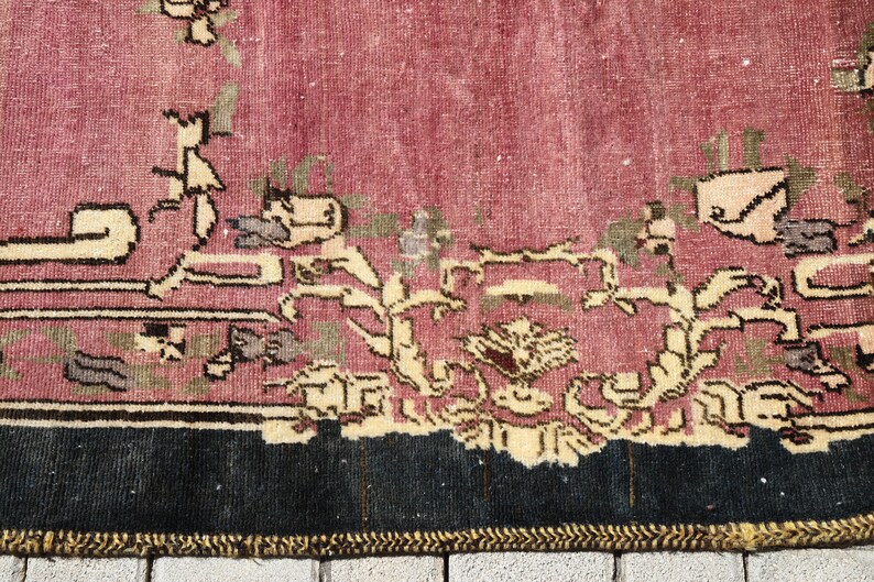 May include: Close-up of the bottom edge of a vintage rug with a pink background and a floral pattern in shades of cream, brown, and green. The rug has a black border and a gold fringe.