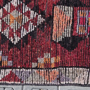 Puede incluir: Primer plano de una alfombra roja, negra y blanca con un borde de flecos. La alfombra tiene un dise&ntilde;o geom&eacute;trico con peque&ntilde;os cuadrados y diamantes.