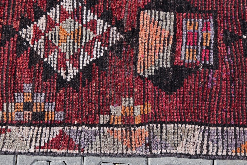 Puede incluir: Primer plano de una alfombra roja, negra y blanca con un borde de flecos. La alfombra tiene un dise&ntilde;o geom&eacute;trico con peque&ntilde;os cuadrados y diamantes.