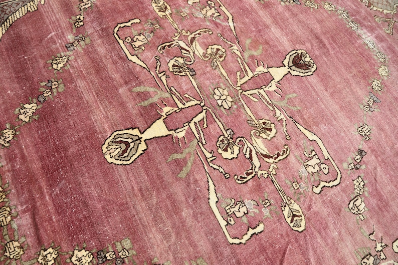 May include: A vintage rug with a pink background and a central design featuring a stylized floral motif in cream and brown. The rug has a border of smaller floral motifs.