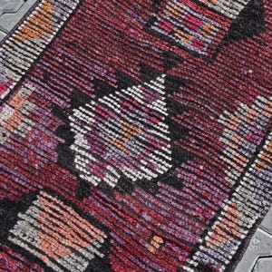 Puede incluir: Una alfombra turca vintage con un patr&oacute;n geom&eacute;trico en tonos rojos, rosas, negros y blancos. La alfombra tiene un aspecto desgastado y est&aacute; hecha de lana.