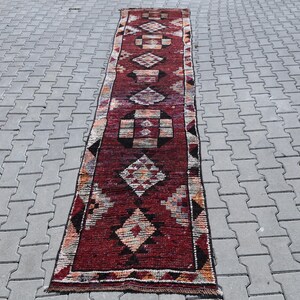 Puede incluir: Una alfombra larga y estrecha de color rojo y crema con un patr&oacute;n geom&eacute;trico. La alfombra tiene un dise&ntilde;o tradicional con formas de diamantes y cuadrados repetidos.