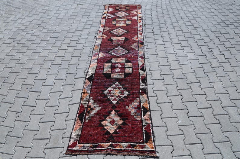 Puede incluir: Una alfombra larga y estrecha de color rojo y crema con un patr&oacute;n geom&eacute;trico. La alfombra tiene un dise&ntilde;o tradicional con formas de diamantes y cuadrados repetidos.