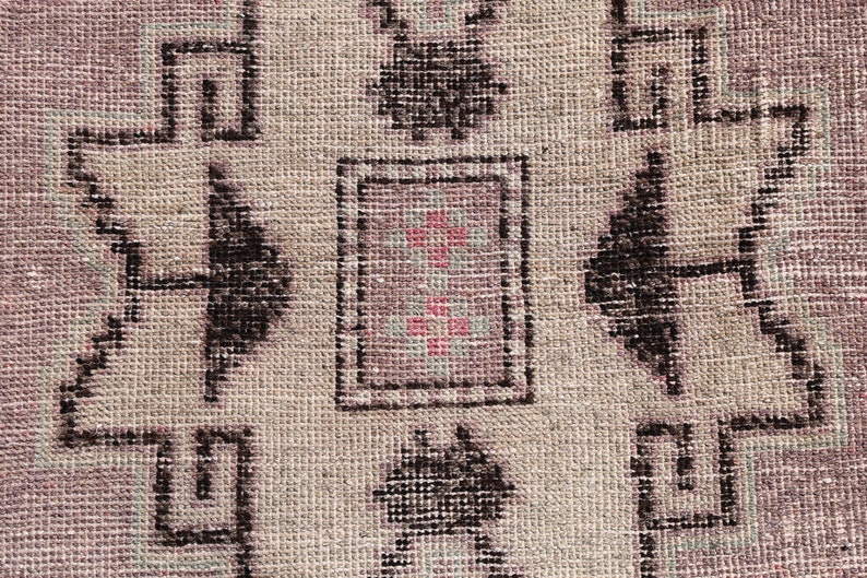 May include: Close-up of a beige rug with a geometric pattern in black and pink. The pattern features a central square with a smaller square inside, surrounded by four larger squares.