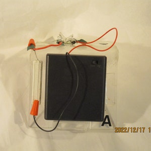 Puede incluir: Un paquete de baterías negro con un cable rojo y un cable negro conectados a él. El paquete de baterías está sobre una base de plástico transparente con una etiqueta blanca que dice "A".