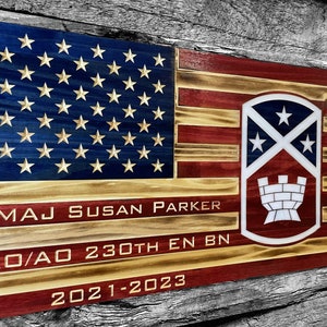 May include: A wooden American flag with a red, white, and blue color scheme. The flag has a distressed look and features a military unit insignia with a castle and stars. The text "MAJ SUSAN PARKER XO/AO 230TH EN BN 2021-2023" is engraved on the flag.
