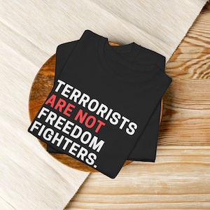 May include: Black t-shirt with the slogan "TERRORISTS ARE NOT FREEDOM FIGHTERS" in white and red. The folded shirt rests on a wooden plate and a cream-colored cloth.