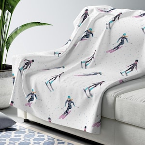 May include: White throw blanket featuring a repeating pattern of skiers in black, teal, and pink. The skiers are depicted in action, with a scattering of small blue and gray dots. The blanket is draped over a light gray sofa.