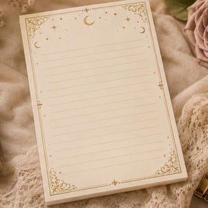 May include: A rectangular notepad with cream-colored paper featuring horizontal lines and gold-toned decorative borders. The design includes celestial motifs such as a crescent moon and stars at the top, with ornate filigree patterns in the corners.
