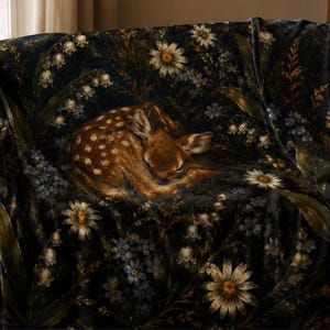 May include: A soft throw blanket featuring a dark, moody background with a printed illustration of a sleeping fawn curled in the center, surrounded by detailed white daisies, blue forget-me-nots, and lily of the valley flowers.