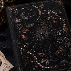 May include: A black hardcover book featuring an intricate cover design of a spider web, a central spider, dark floral motifs, and a curved string of pearls. The cover includes ornate gold-toned corner accents and subtle metallic highlights.