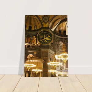 May include: A close-up view of the interior of a mosque with ornate gold chandeliers hanging from the ceiling. The ceiling is decorated with intricate patterns and the walls are a warm brown color. A large, circular plaque with Arabic calligraphy is visible in the center of the image.