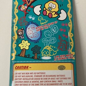 K&ouml;nnte beinhalten: Eine t&uuml;rkisblaue und gelbe Pappverpackung f&uuml;r ein Dinkie Dino Spielzeug. Die Verpackung zeigt einen Comic-Dinosaurier mit einem Schnuller und dem Text "Sieh zu, wie ich Tag f&uuml;r Tag gr&ouml;&szlig;er werde!" und "Ich bin sehr gl&uuml;cklich zur Badezeit!" Die Verpackung enth&auml;lt auch ein Warnschild mit Anweisungen zur Verwendung von Batterien.