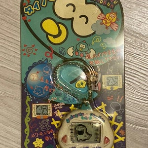 May include: A white and blue digital pet keychain with a screen displaying a cartoon character. The keychain is packaged in a plastic blister pack with Japanese text and illustrations. The text on the packaging reads "Rakuraku Dinokun" and "ITEM NO.: TK-910".