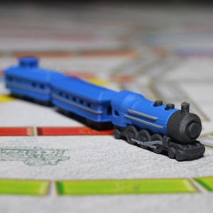 May include: A blue toy train with a black smokestack and wheels sits on a game board. The train is composed of an engine and two cars. The game board has colored lines and a map design.