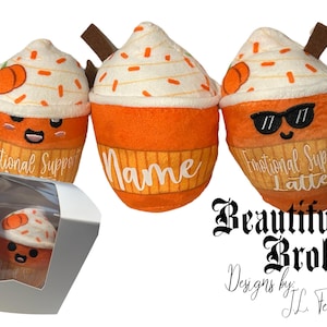May include: Three plush orange and white pumpkin spice latte-themed cupcakes with embroidered faces and text. The cupcakes are labeled "Emotional Support Latte", "Name", and "Emotional Support Latte". The cupcakes are sitting on a white surface with a white box in the foreground.