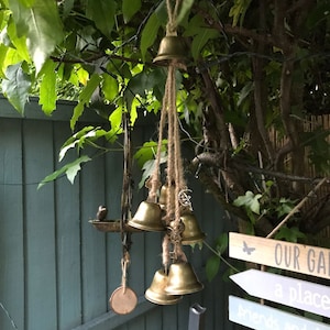 May include: A wind chime with multiple gold bells hanging from a tree branch. The bells are attached to a string with a wooden bead. The wind chime is hanging in front of a blue wooden fence. The sign reads "Our Garden, a place for friends and family, and love."