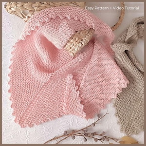 Può includere: Uno scialle rosa chiaro lavorato a maglia con bordo smerlato è drappeggiato su un cestino intrecciato. Una sciarpa beige lavorata a maglia è visibile a destra. Il testo "Easy Pattern + Video Tutorial" è in alto a destra. Lo scialle è realizzato con filato morbido e strutturato.