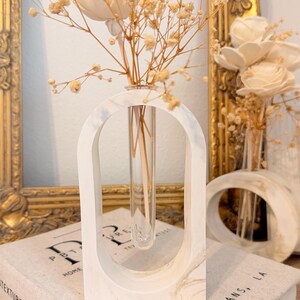 May include: A white marble-patterned arch-shaped frame holding a clear glass test tube vase. The vase contains dried beige baby's breath flowers. The frame sits on a book with text that includes "NEW ORLEANS, LA" and "TEXTURES".
