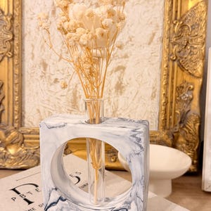Puede incluir: Un florero cuadrado de resina con efecto mármol blanco y gris que presenta un recorte circular central que sostiene un tubo de ensayo de vidrio transparente. El tubo contiene un ramo de flores secas color crema con tallos finos y claros.