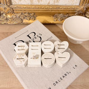 May include: A collection of seven off-white ring holders with a marbled pattern, featuring various geometric shapes including hexagons, rectangles, circles, and hearts, each with a horizontal slot for rings, with one gold ring displayed.